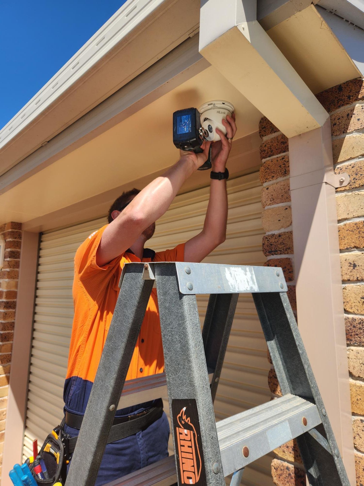 Technician in an orange shirt installing a security camera under the eaves of a Dalby home